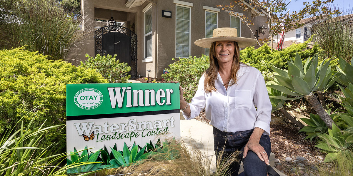 Landscape Contest Winner kneeling by a winner's sign in her yard